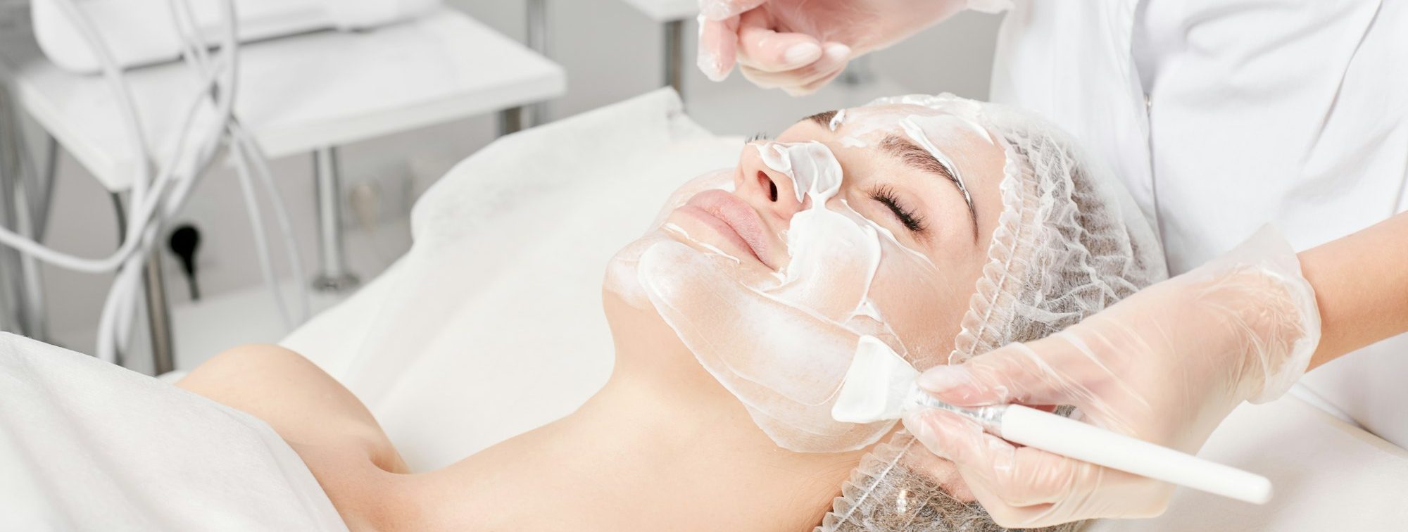 Beautician applies cream mask on woman face for rejuvenation face skin, procedure in beauty salon